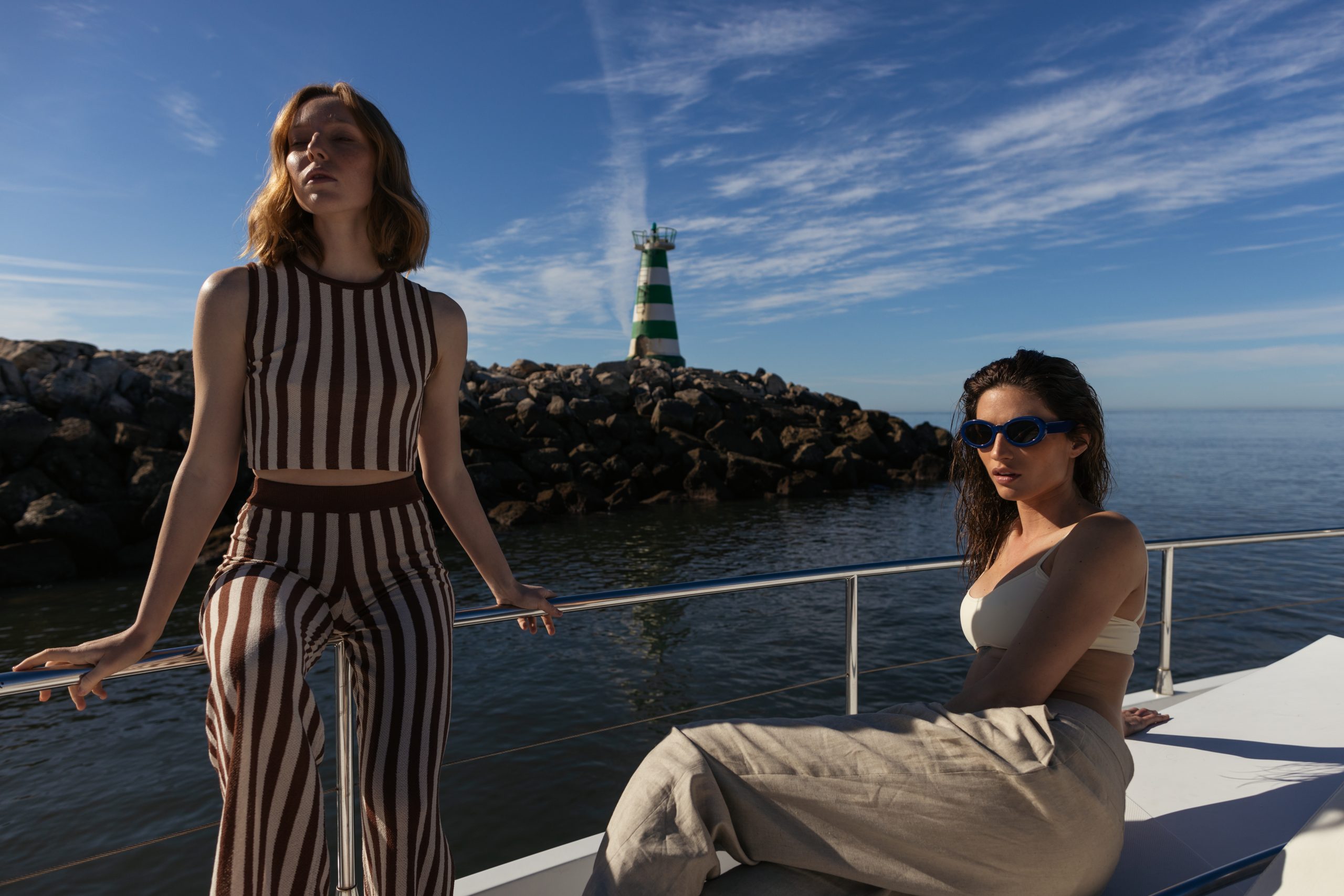 Two fashionable women enjoying a sunny day on a yacht, with a scenic view of the ocean and a green-and-white striped lighthouse on a rocky outcrop in the background. The clear blue sky and reflective water create a luxurious and serene atmosphere.