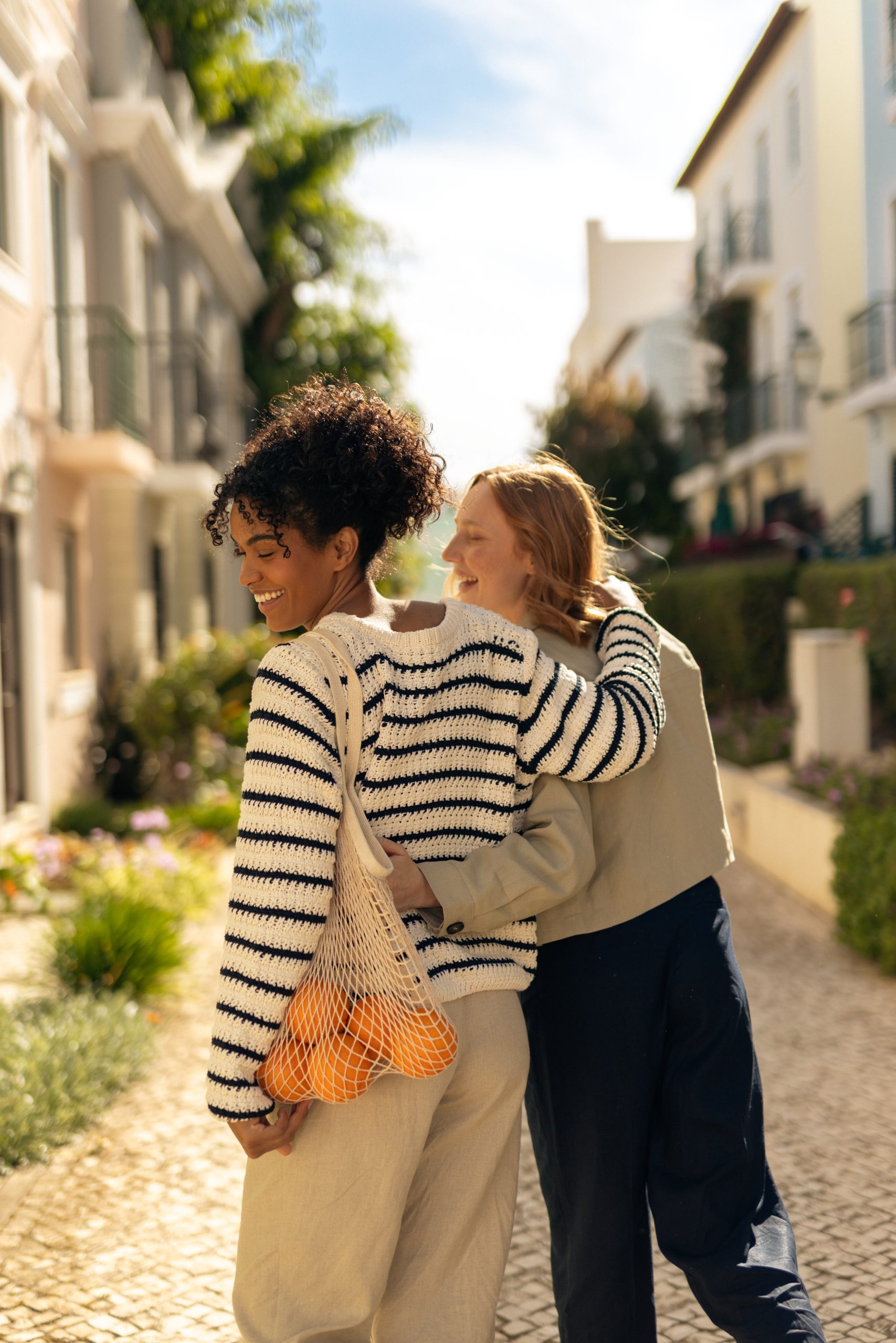 Two people walking down a sunlit, narrow street lined with buildings and greenery. One person has an arm around the other, and they are carrying a mesh bag with oranges.