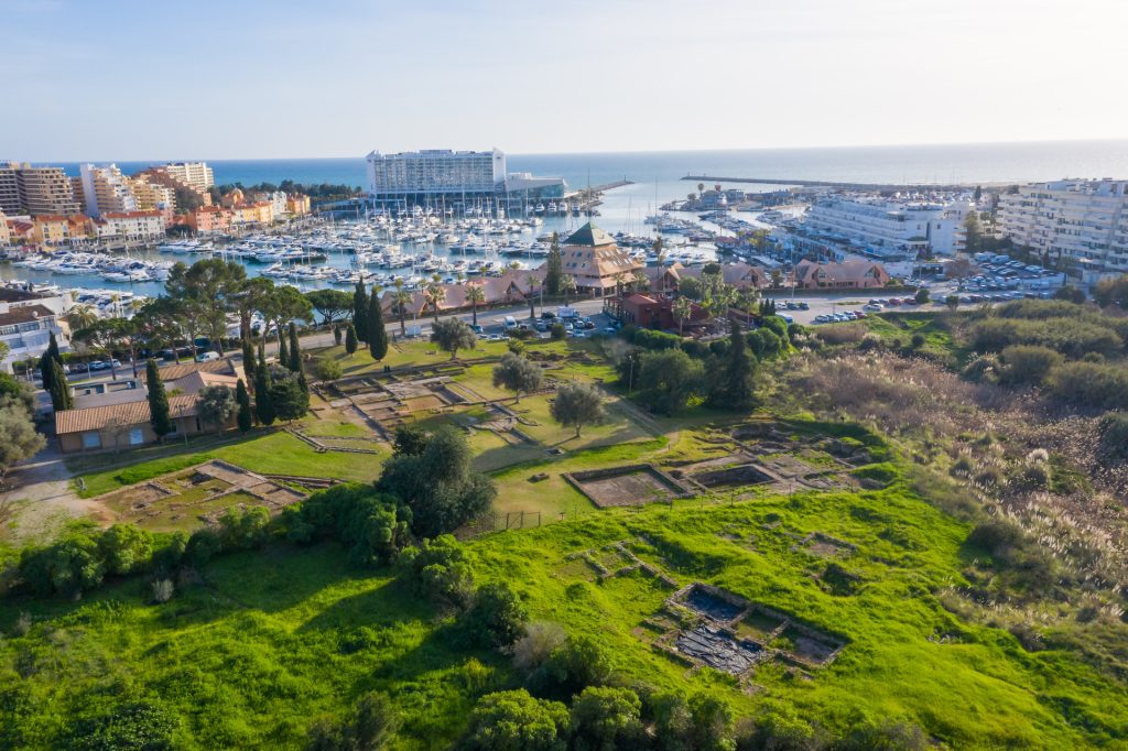 Aerial view of Vilamoura, a coastal town in the Algarve, Portugal, featuring a marina filled with boats, green areas with archaeological ruins, and buildings in the background with the sea visible beyond the marina.