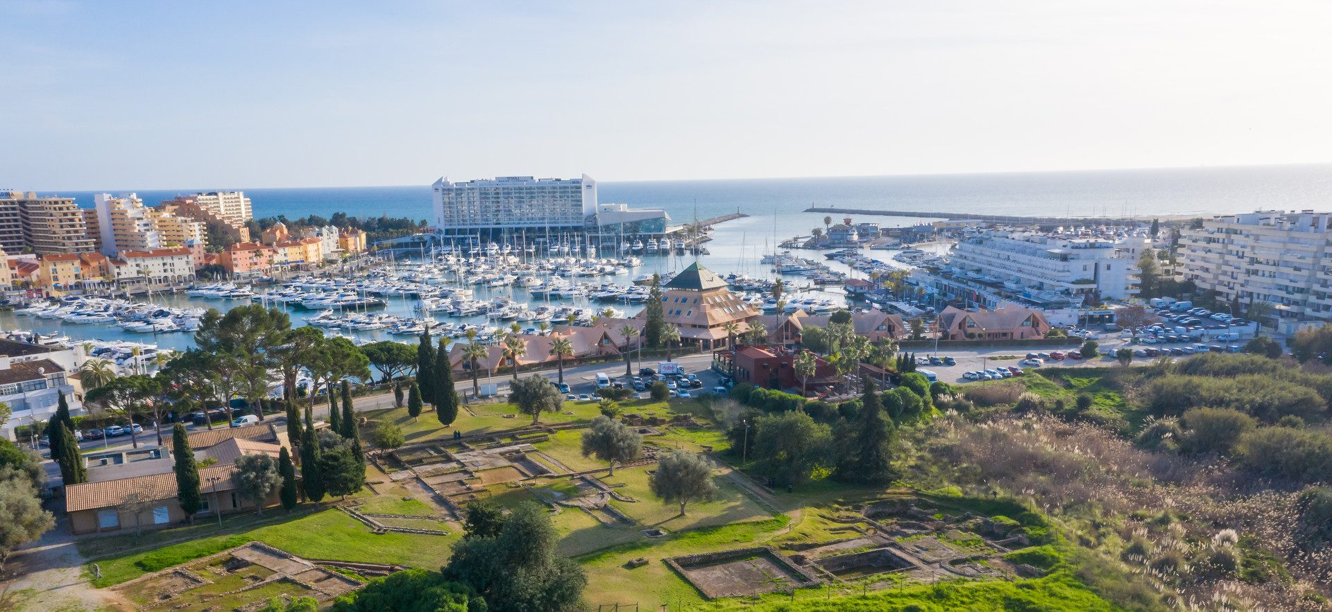 Aerial view of Vilamoura, a coastal town in the Algarve, Portugal, featuring a marina filled with boats, green areas with archaeological ruins, and buildings in the background with the sea visible beyond the marina.